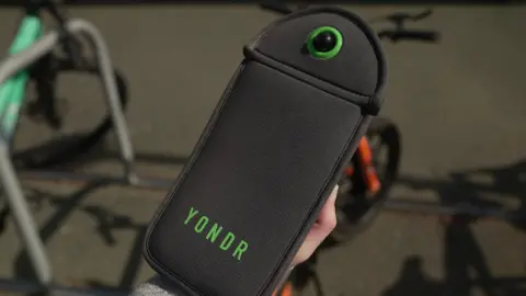 The photo shows a black pouch, which is being held in front of various bikes which are locked up. The pouch reads YONDR in green font. 