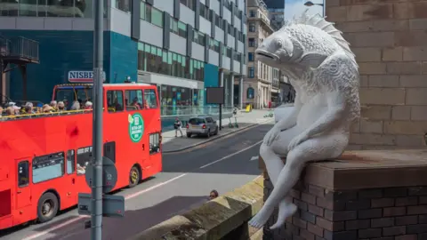 Chloe Lloyd The sculpture Skalli in situated on the end of the plinth above the street below. A red open top tourist bus is passing by.
