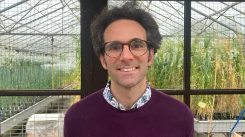 Owen Sennitt/BBC A man wearing glasses, a maroon jumper and shirt smiles towards the camera in front of a glasshouse where wheat is being grown for experiments