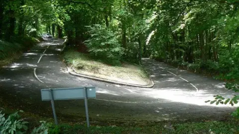 Colin Madge / Geograph A hairpin bend on a steep hill surrounded by mature trees in leaf. 