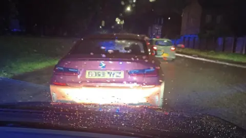 The back of a red car with the number plate blurred. The car is on the side of the road at night.