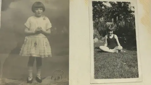 David Turner Two old black-and-white photographs of a young girl with dark bobbed hair, side-by-side in an album. In one picture she is standing with her hands clasped and is wearing a white frilly dress. In the other she is wearing what looks like a black bathing suit and is sitting on some grass.