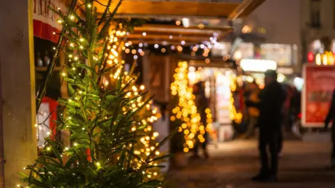 A Christmas tree outside, with lights on it. A high street scene behind it.