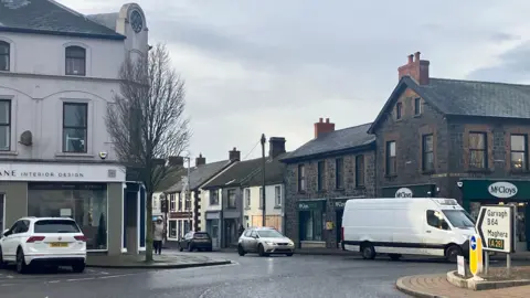 The centre of Kilrea, showing a small roundabout, grey buildings and a number of white vehicles.