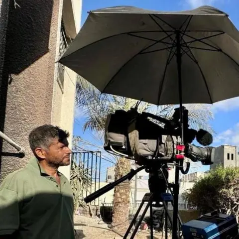 Reuters A man stands to the side of a TV camera watching it as he films. He is stood under an umbrella in Gaza
