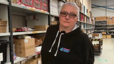 A woman with short white hair looking into the camera. She is not smiling. She is wearing a black jumper with white strings. There are shelves full of boxes behind her. 