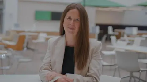 Wiltshire Council Lucy Townsend smiles at the camera. She has long dark hair, is wearing a beige jacket and sites in an empty cafe area
