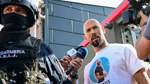 Andrew Tate looks at the camera while being brought in by police, a masked police officer wearing a helmet looks on while reporters gather round Tate, taken outside the Tate brothers' residence in Bucharest on Wednesday night