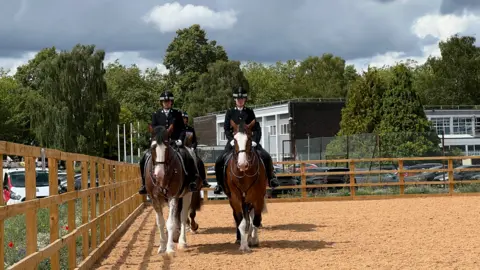 Mounted police officers riding horses side by side in a fenced outdoor riding arena, with trees, buildings and dark clouds in the background.