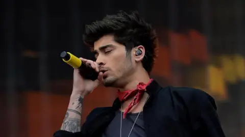 PA Media Zayn Malik of performing on stage. He has his eyes closed as he sings into a microphone. He is wearing a red neckerchief with a black T shirt and jacket.
