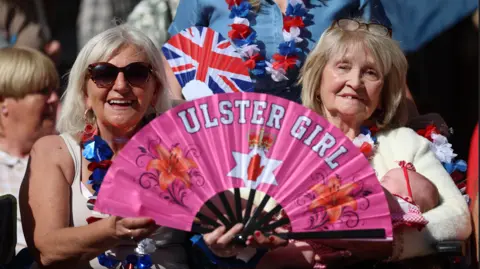 Two women with blond hair are holding up a pink fan that says Ulster girl. One is holding a baby. A crowd is behind them. 