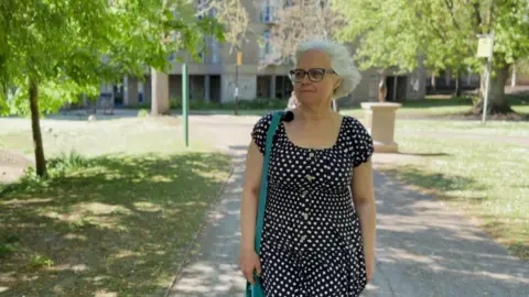 A woman in a black dress with white polka dots walking along a shaded park path, carrying a turquoise shoulder bag, with trees, grass, a pond and a multi‑windowed building in the background on a sunny day.