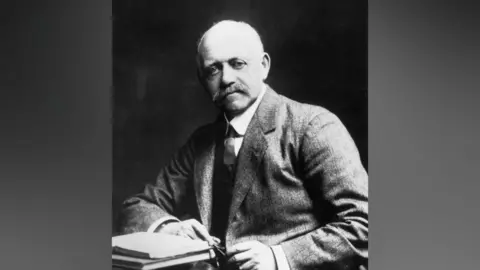 A black and white photo of a middle-aged man wearing a suit. There are books ladened on a table next to him.