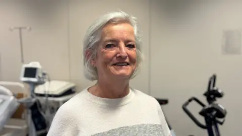 Anne Smith (grey hair, white and grey round necked top) smiling and looking at the camera. Monitoring equipment and the handles of exercise bikes in the background.