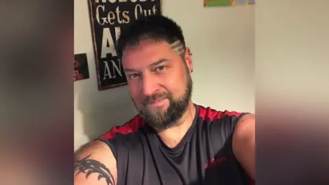 Sean is in his 40s with dark hair and beard looking directly at the camera. He is wearing a red and black top and has a tattoo on his right arm.