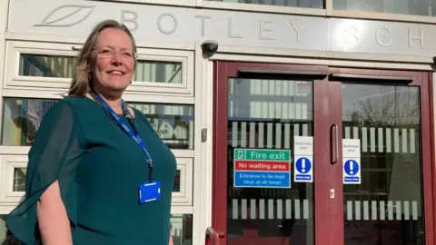 A woman is standing outside a school entrance. She is wearing glasses and a green top and looks very proud.