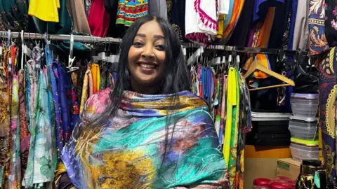 BBC Athar Alsadek wearing colourful clothing. She is in a market and there are clothes on a rail behind her. She has long black hair and is wearing a smile. 