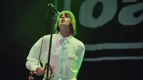 Getty Images A blurry image shows Liam Gallagher on stage at Knebworth, singing into a microphone on a tall stand and holding a tambourine