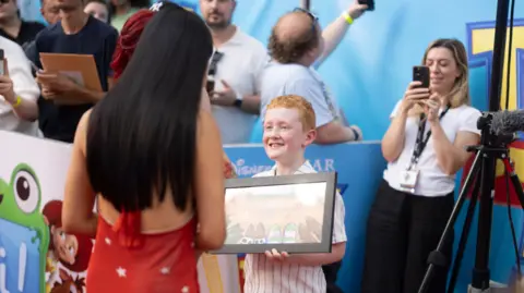 BBFC A young boy is smiling while holding a framed copy of his winning design. A woman with a red dress with white stars and long black hair is talking to him. Several people can be seen behind, including a woman who is holding a mobile phone.