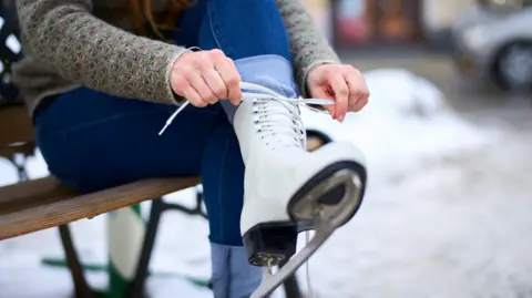 A stock image of a woman tying a lace of an ice skate while sat down on a wooden bench.