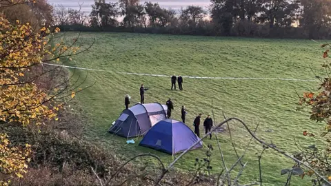 BBC/Paul Murphy Two tents and a cordon on a greenfield with trees and woodland surrounding it. There are eight police officers in uniform standing near the tents