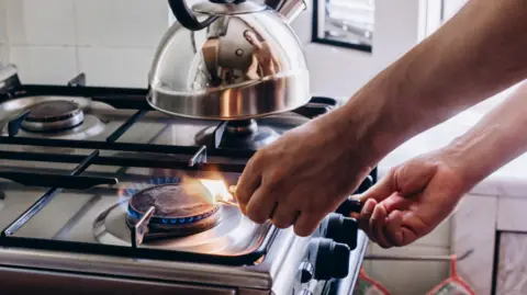 A person lights a gas stove using a match. There is a silver kettle sitting on one of the rings.