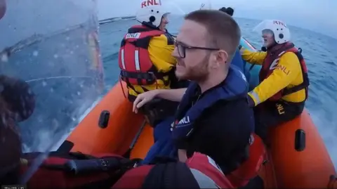 A man with short hair and glasses sits in a lifeboat at sea as he is rescued by RNLI volunteers. 