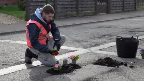 A man with short dark hair and beard wearing a pink high visibility vest plants flowers in a pothole on a road. 
