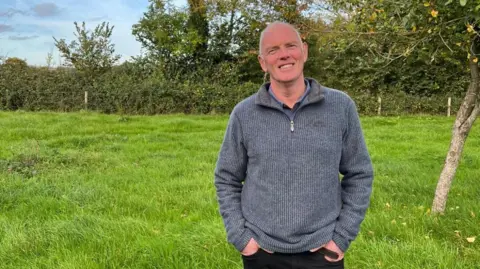 Mark Humphrey A man is wearing a blue quarter zip jumper and smiling at the camera. He's stood in a grassy field, with trees and hedgerows behind him.
