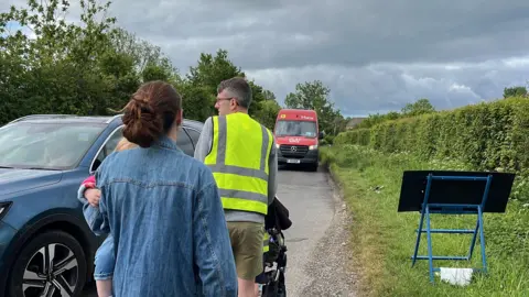 George Carden/BBC Two people with a pram and holding a baby walk along the country lane which has hedges and a grass verge but no footpath. There's cars and vans queuing along the road and one of the people is wearing a high vis vest