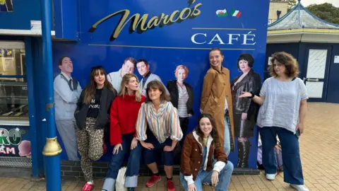 IJPR Five women are smiling while standing in front of the blue Marc's Cafe sign at Barry Island, with one woman kneeling on the floor. The sign includes photos of some of the cast of Gavin and Stacey, including Joanna Page as Stacey and Rob Bryden as Uncle Bryn. Ruth Jones is on the right, standing next to a photo of her as Ness.