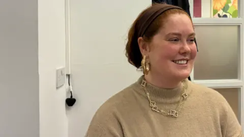 Zavier is smiling and has red hair which is tied up in a bun, has rosy cheeks and is wearing a gold jumper with gold jewellery