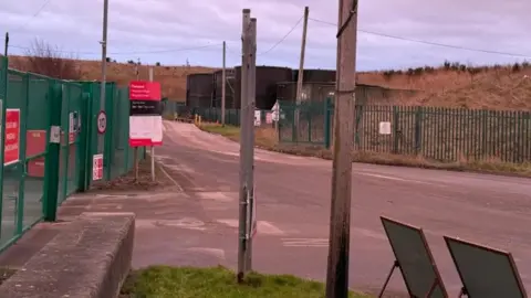 The entrance to the landfill site is surrounded by green fences with red and white signs attached.