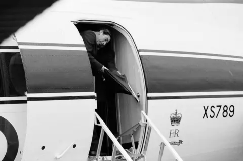 Getty Images A black-and-white image of a man standing in the doorway of a black and white XS789 plane with royal insignia of a crown and the lettering E II R.