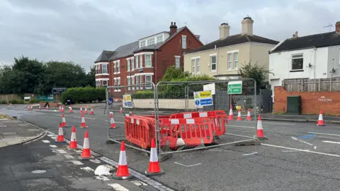 A section of a road is surrounded by both grey metal fencing and bright orange plastic barriers. There are signs on it that read: "Danger. Keep Out." White and orange cones line the road beyond it and there are houses bordering the road on one side.