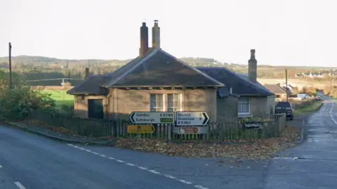 Google Two roads meet on a corner. A house in situated on the corner, with a sign pointing left for Gordon and Edinburgh and right for Stichill and Greenlaw. It is a countryside setting, with green fields and bushes in the background