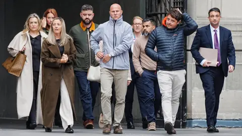 PA Media A group of nine people, including four women and five men, cross a stone square with sombre, determined expressions. 