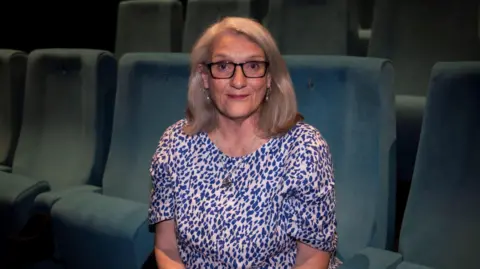 Kat Gollock Ginnie Atkinson, with blonde shoulder length hair and glasses, and wearing a purple patterned dress, sits in the refurbished cinema boasting pristeen blue velvet seating