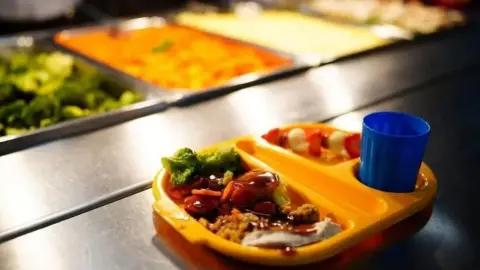 A school meal in a yellow plastic tray. The tray is sectioned into three with the main meal on the left, a dessert in the top right corner and a blue drinks cup in a circular holder on the bottom right. It stands on a stainless steel counter with trays of food in the distance.