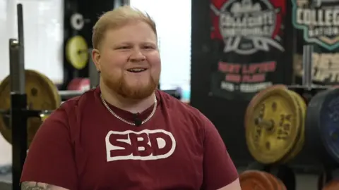 BBC Ray Bowring sitting smiling, wearing a burgundy t-shirt with the letters SBD, in front of racks of weights in a gym