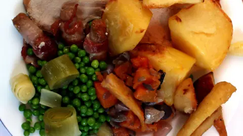 A plate is filled with Christmas dinner and all the trimmings. Turkey can barely be seen under crisply roasted potatoes, roasted parsnips, carrots, with peas and chopped leeks and pigs in blankets which are sausages wrapped in bacon.