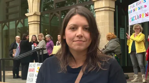 Local Democracy Reporting Service Lauren Bunting, co-founder of West Northants SEND Action Group, stands on the steps of Northampton Guildhall, among other protesters holding up signs and placards. She has brown hair, which is slightly longer than shoulder-length, and is wearing a dark-coloured top.