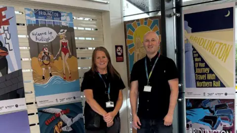 Northern Two adults stand beside a wall of colourful railway posters