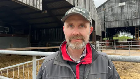 Photograph of Chatham looking into the camera with a neutral expression. He has a short, dark grey beard. He is wearing a red and black waterproof jacket with a grey overall over the top. Chatham is also wearing a dark green cap with 'McHale' written on it in white lettering. He is stood in front of two huge barns, clad in corrugated iron. There are metal gates and fences in front of the entrances, with straw strewn across the floors. 