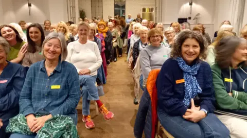 BBC A large group of WI members are seated in a room with a walkway through the middle. They are all smiling and looking straight at the camera. 