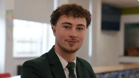 Tom is wearing a green blazer and a green tie with a white shirt. He has brown hair and is in the school canteen with tables, a white wall and large windows behind him.