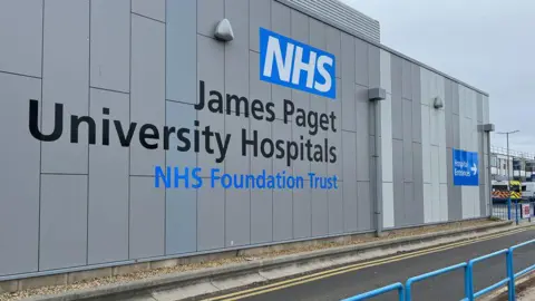 A grey wall of a hospital which says NHS James Paget University Hospitals and in front of it a road. Beyond it to the far right can be seen two ambulances