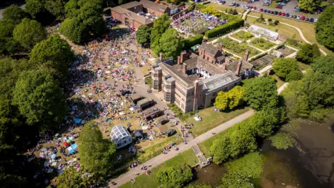 Aerial view of a large stately home building and its gardens - with many people in the gardens below with parasols and picnic blankets.