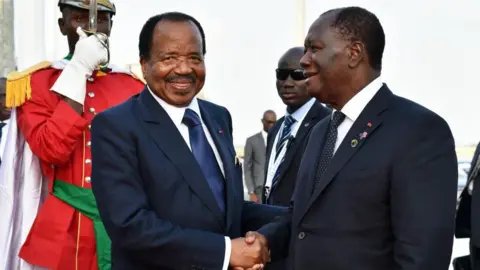 AFP/Getty Images Ivory Coast's President Alassane Ouattara (R) in a dark grey suit and tie welcomes a smiling Cameroon's President Paul Biya, wearing a navy suit and tie, at Félix-Houphouët-Boigny International Airport in Abidjan on 28 November 2017 a couple of days ahead of a African Union-European Union summit. Behind Biya to the left can be seen a guard in a red buttoned coat with epaulets, green sash, white gloves who hold up a ceremonial sword vertically in front of his face. A suited man in dark glasses and wearing a lanyard stands behind Ouattara and stares into the camera.