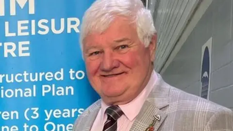 Amber Valley Borough Council A man with white hair, smiling and wearing a grey chequed suit and pink shirt with a dark striped tie.